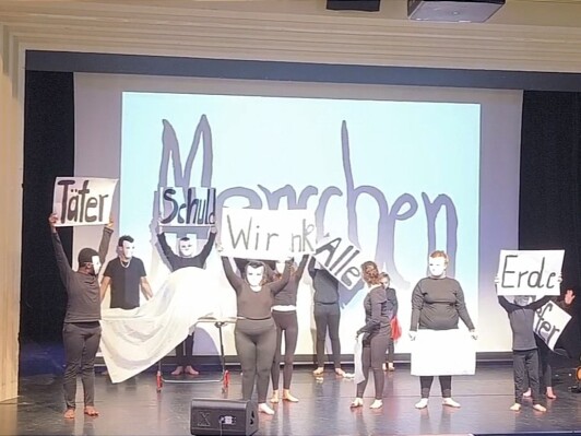 Theatergruppe auf der Bühne mit weißen Masken und Schildern