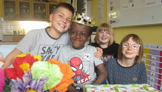 4 Schüler stehen zusammen, ein Kind hat eine Krone auf. Vor den Kindern steht ein Blumenstrauß aus Papier