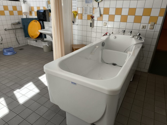 Therapiebadewanne in der Mitte und Waschbecken, Dusche und Sitzgelegenheit, Tritthocker